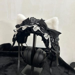 Puede incluir: Una diadema de orejas de gato en blanco y negro con encaje y un lazo. La diadema tiene orejas blancas y esponjosas y está decorada con encaje negro y un lazo negro. La diadema se muestra con auriculares negros.