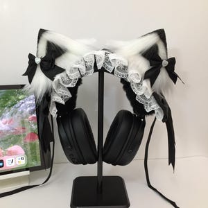 May include: Black and white faux fur cat ears attached to black over-ear headphones. The ears feature white lace trim, black satin bows with small silver heart charms, and long black ribbons hanging down from the sides of the headset.