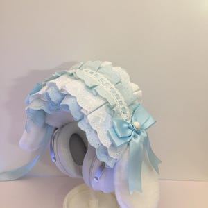 Puede incluir: Una funda para auriculares con orejas de conejo azul claro y blanco con un diseño con volantes. La funda tiene un lazo azul claro y orejas blancas y esponjosas. Los auriculares son blancos y están sobre un soporte blanco.