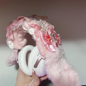 Könnte beinhalten: Weiße Kopfhörer, verziert mit rosa Kunstfell, Spitze und einer Satinschleife mit einem geschmückten Mittelstück. Die Kopfhörer haben einen flauschigen, hellrosa Schwanz und ein dekoratives Stirnband mit Schichten aus rosa und weißer Spitze.