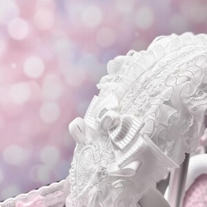 May include: White lace bonnet with a large bow and delicate ribbon details. The bonnet features intricate embroidery and layered ruffles, creating a romantic and vintage aesthetic. The background is a soft, blurred mix of pink and purple.