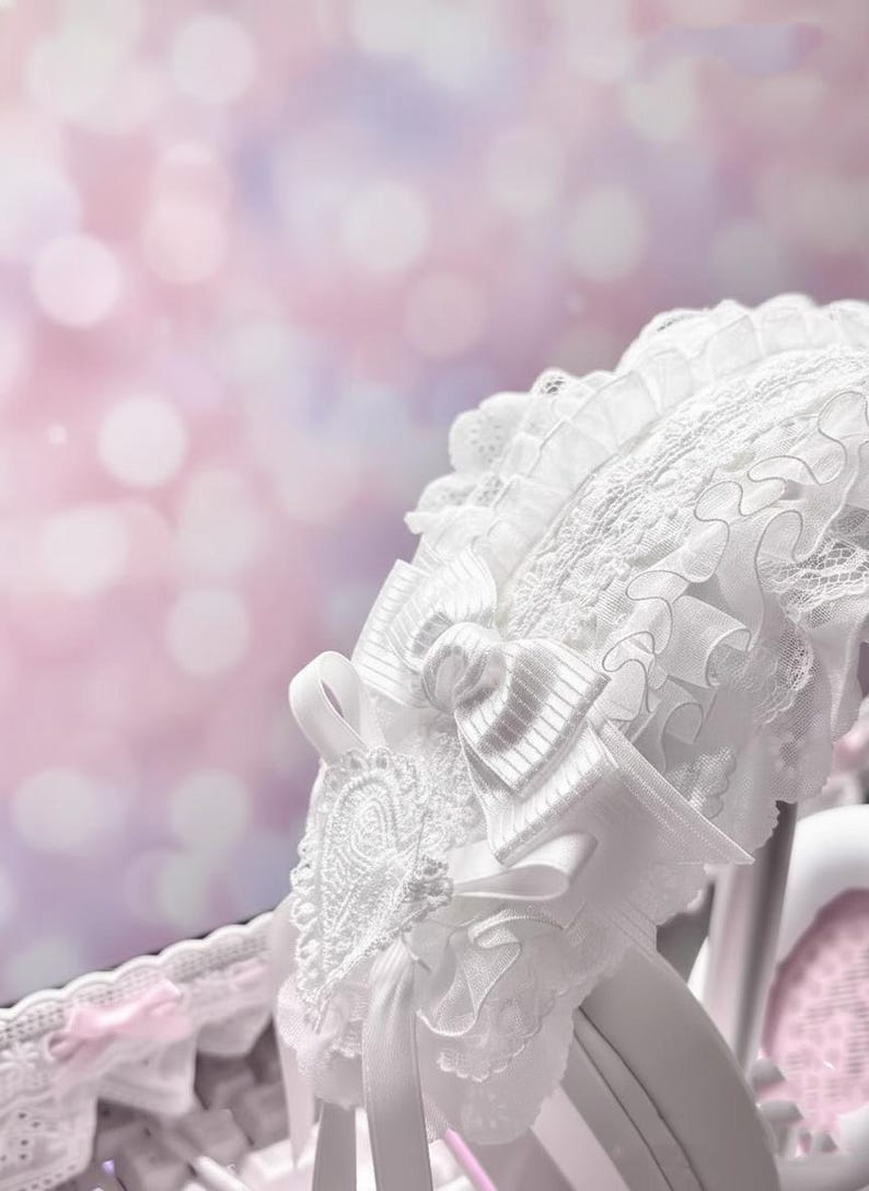 May include: White lace bonnet with a large bow and delicate ribbon details. The bonnet features intricate embroidery and layered ruffles, creating a romantic and vintage aesthetic. The background is a soft, blurred mix of pink and purple.