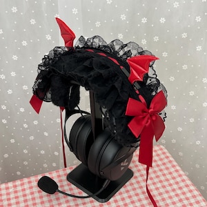 Puede incluir: Una diadema negra con cuernos y alas de diablo rojos, adornada con encaje negro y un lazo rojo. La diadema se exhibe en un soporte negro para auriculares, con auriculares negros. El fondo tiene un patrón floral blanco.