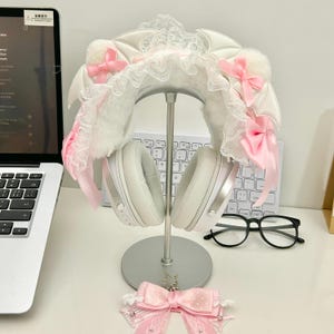 Puede incluir: Auriculares inalámbricos blancos con una funda decorativa para la diadema con encaje blanco, piel sintética blanca, pequeñas alas de murciélago blancas y lazos de satén rosa claro. Un accesorio de lazo rosa claro con lunares y estrella.