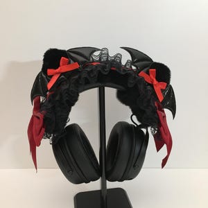 Puede incluir: Auriculares negros con una diadema decorativa con alas de murciélago negras, lazos rojos y encaje negro. Los auriculares se exhiben en un soporte negro sobre un fondo blanco. La diadema añade una estética gótica o cosplay.