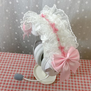 Puede incluir: Auriculares para juegos blancos con una diadema decorativa de encaje y lazo rosa. La diadema presenta encaje blanco, pequeños lazos rosas y una cinta rosa. Los auriculares están sobre un soporte blanco, con un micrófono gris.