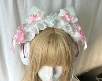 May include: A white plush cat ear headband featuring white lace trim and multiple light pink satin bows. The headband is worn over white headphones with a microphone attachment, positioned above blonde hair with straight-cut bangs.