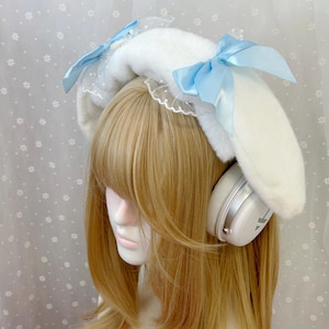 Może przedstawiać: Biała pluszowa opaska z długimi, miękkimi, klapniętymi uszami, ozdobiona delikatną białą koronką i dwiema jasnoniebieskimi satynowymi wstążkami zawiązanymi w kokardy po obu stronach, założona na białe słuchawki nauszne ze srebrnymi akcentami.