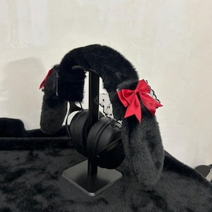 Puede incluir: Auriculares negros con orejas de conejo de piel sintética, lazos de satén rojo y detalles de encaje negro. Los auriculares están sobre un soporte negro, sobre una superficie negra texturizada.