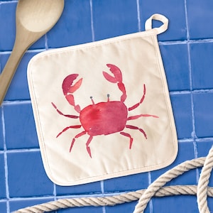 May include: A white cotton pot holder with a red watercolor crab design. The crab is facing right and has a light pink body with dark red legs and claws.