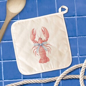 May include: Off-white square pot holder with a red lobster illustration wearing a blue and white striped bow. The pot holder has a white loop for hanging. A wooden spoon and rope are in the background.
