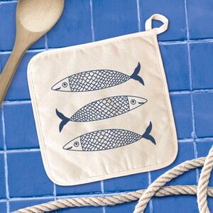 May include: A beige pot holder with a loop for hanging, featuring three stylized blue fish. The fish have detailed scales and dark blue fins. The background is a blue tiled surface, with a wooden spoon and rope visible.