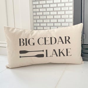 May include: A rectangular beige pillow with the words "BIG CEDAR LAKE" printed in black, along with two black oars. The pillow is made of a soft fabric and is likely used for home decor.