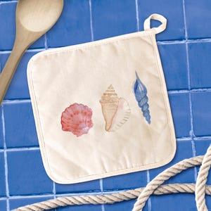 May include: Off-white square pot holder featuring watercolor illustrations of three seashells in shades of pink, tan, and blue. The pot holder has a loop for hanging. A wooden spoon and rope are in the background.
