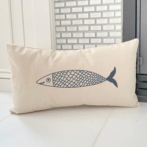 May include: A rectangular beige pillow with a navy blue fish illustration. The fish has a detailed scale pattern and a curved tail. The pillow is likely made of a natural fabric, and is a decorative item for a home.