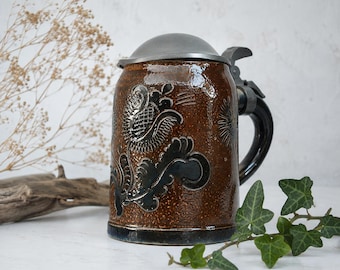 Puede incluir: Una jarra de cerveza de cerámica marrón con un acabado texturizado y moteado, y una tapa de metal oscuro. El cuerpo presenta un relieve floral oscuro. Incluye un asa curva resistente y un mecanismo de tapa de metal con bisagras en la parte superior.