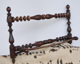 Könnte beinhalten: Ein dunkelbraunes Wandregal aus Holz mit zwei horizontalen Stangen und dekorativen gedrechselten Spindelelementen. Das Regal ist über einem cremefarbenen Stoffstück angebracht, das eine schwarze handschriftliche Aufschrift auf der Oberfläche zeigt.