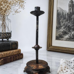 May include: A tall, dark, weathered metal candlestick holder featuring a slender central rod, a small circular drip plate, and a wider, flat circular base supported by three small feet. The metal surface shows signs of oxidation and dark patina.