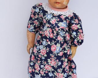 May include: An antique doll with a painted face and a floral print dress. The dress is navy blue with pink and blue flowers, a white lace collar, and short sleeves. The doll is standing on a wooden base.