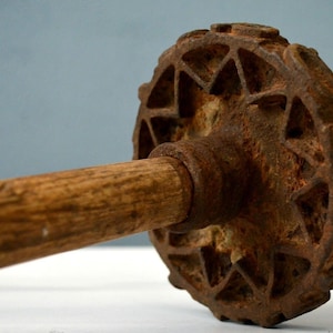 May include: An antique, rusty metal butter churn with a wooden handle. The churn's head has a geometric design. The handle is a light brown color. The churn is set against a light blue background.