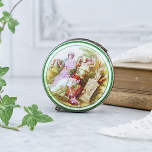 May include: A round metal trinket box featuring a colorful painted scene of two figures in period clothing sitting near a stone pedestal. The lid is framed by a thin green border and a metallic edge, showcasing a detailed illustration of people.