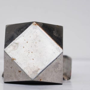 Op de afbeelding: Close-up van een paar vierkante metalen manchetknopen. Elke manchetknop heeft een zilveren ruitvorm in een donkerder, mogelijk geoxideerd, metalen vierkant. Het ontwerp is eenvoudig en geometrisch.