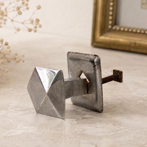 May include: A silver-toned metal cabinet knob featuring a faceted, pyramid-shaped front and a square backplate. The hardware includes a threaded metal bolt extending from the rear, showing signs of oxidation and a weathered, metallic finish.