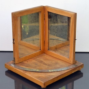 Op de afbeelding: Een vintage houten hoekspiegel met twee scharnierende panelen. De spiegel staat op een houten voet met een gebogen metalen detail. Het frame en de voet hebben een warme, natuurlijke houttint en de spiegels vertonen tekenen van ouderdom.