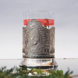May include: Silver-plated glass holder with an ornate design and red stripes around the glass. The holder has a base and a decorative top.