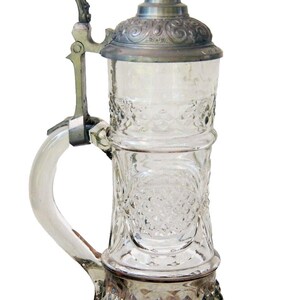 Könnte beinhalten: Klarer Bierkrug aus Glas mit einem dekorativen geätzten Muster. Der Krug hat einen Klappdeckel und einen Griff aus Metall. Der Boden und der Deckel sind silberfarben, und der Griff ist aus klarem Glas.