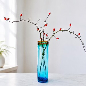 Pode incluir: Um vaso cilíndrico alto de vidro com um gradiente de cor que transita do azul profundo na base para um turquesa mais claro e uma borda âmbar. Dentro do vaso estão ramos finos castanho-escuros adornados com pequenos frutos vermelhos.