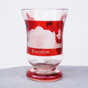 Könnte beinhalten: Ein klares Glas mit rotem Band und Boden. Das Glas hat ein weiß geätztes Design eines Gebäudes und Bäume, mit dem Wort "Pavillon" in weißen Buchstaben. Das Glas hat eine ausgestellte Form und einen abgerundeten Boden.