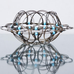 May include: A spherical, openwork metal basket with a complex, interwoven design. The dark metal is accented with small, light blue beads. The basket is reflected in the shiny surface below.
