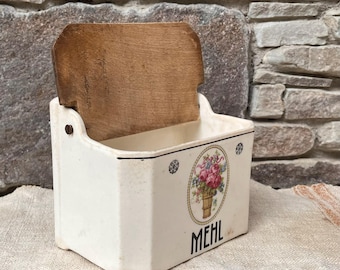 Puede incluir: Contenedor de cerámica color crema con tapa de madera. El contenedor presenta un diseño floral en un marco ovalado y la palabra "MEHL" en negro. Probablemente para almacenar harina u otros productos secos.