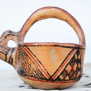 May include: A hand-painted ceramic vessel featuring a rounded bowl, a high arched handle, and a protruding spout. The exterior is decorated with geometric patterns in black and red paint over a tan base, including cross-hatching and diamond shapes.