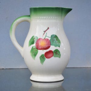 Pode incluir: Um jarro de cerâmica branca com uma borda verde e um desenho pintado de maçãs vermelhas e folhas verdes.