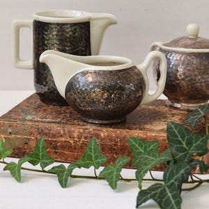 Könnte beinhalten: Ein Set aus drei vintage Keramik-Milchkännchen, Zuckerdose und Kanne mit gehämmertem Metalloberflächenfinish. Die Teile sind braun und schwarz mit einem weißen Rand. Das Set wird auf einem braunen Buch mit grünem Efeu gezeigt.