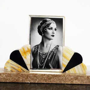May include: Art Deco style photo frame with a black and white portrait. The frame has a rectangular photo area and a fan-shaped design with black, yellow, and white accents. The base is a mottled brown color.