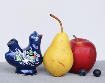 May include: A still life composition featuring a blue ceramic bird whistle with floral accents, a yellow pear, and a red apple. Two small, dark berries are also present. The objects are arranged on a gray surface.