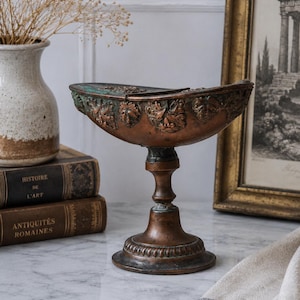 May include: A bronze-toned metal pedestal bowl featuring a wide, shallow basin with a decorative relief of leaves and faces around the exterior rim. The bowl is supported by a slender, tiered stem and a circular base with a ribbed edge detail.