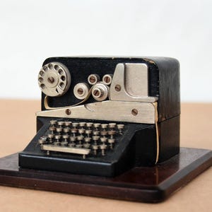 Könnte beinhalten: Eine Miniatur-Vintage-Schreibmaschine mit einem schwarzen Holzgehäuse und silberfarbenen Metallkomponenten. Sie steht auf einem dunkelbraunen Holzsockel. Die Tasten sind sichtbar, und das Design erinnert an die Technologie des frühen 20. Jahrhunderts.