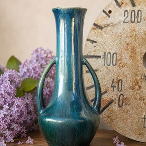 Peut inclure: Un vase en céramique haut et élancé avec un vernis brillant bleu sarcelle profond. Le vase présente un col étroit, une base arrondie et deux anses incurvées symétriques s'étendant du col jusqu'à l'épaule du récipient.