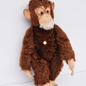 May include: A brown plush monkey with a light tan face, ears, hands, and feet. The monkey has red eyes, a white beard, and a small gold tag hanging from its neck. The monkey is standing upright.