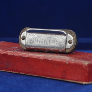 May include: A silver Hohner harmonica sits on a red rectangular box. The harmonica features the engraved brand name "Hohner". The box is a deep red colour, set against a dark blue background.