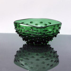 May include: A green glass bowl with a textured, bumpy surface. The bowl is square shaped and has a slightly rounded bottom.