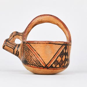 May include: A small, hand-formed terracotta vessel with a high arched handle and a protruding spout. The exterior surface features hand-painted geometric patterns in black and dark red, including cross-hatching and diamond shapes on a tan base.
