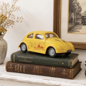 May include: A yellow toy car with a rounded body, grey wheels, and the text "AL SZERVIZ" printed on the side. The car features a black front license plate with white text and is positioned on top of two stacked vintage hardcover books.