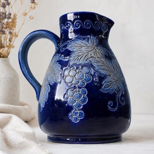Puede incluir: Una jarra de cerámica en color azul cobalto profundo con acabado brillante y un diseño en relieve de racimos de uvas y hojas de parra. El recipiente cuenta con un asa curva, una base ancha y un patrón decorativo de volutas en el borde.