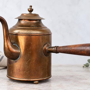 May include: A cylindrical copper teapot featuring a long, curved spout, a removable lid with a small finial, and a horizontal wooden handle. The metal body displays a weathered patina with visible surface scratches and rests on small feet.