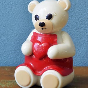 May include: A white teddy bear figurine with a red heart-shaped apron. The bear has black eyes, a tan nose, and tan inner ears. The bear is sitting with its legs extended and has tan-colored feet.